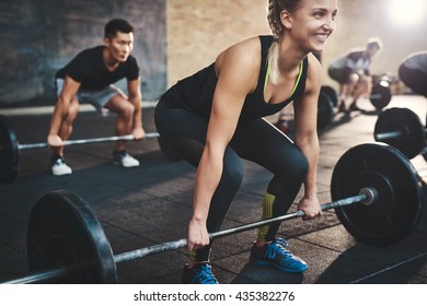 Cheerful muscular young woman with ponytail and black tights performing dead lift barbell exercises with other students
