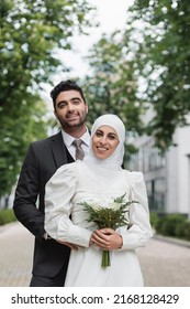 cheerful muslim bride in wedding hijab and white dress holding bouquet near groom outside