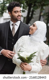 cheerful muslim bride in wedding hijab and white dress holding bouquet and looking at  groom outside