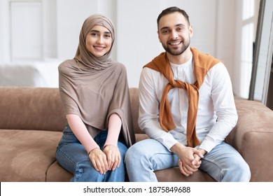 Cheerful Muslim Couple Sitting On Sofa Smiling To Camera In Living Room At Home. Portrait Of Modern Arab Family Of Two, Happy Young Husband And Wife In Hijab Posing Together Indoor Concept
