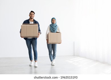 Cheerful Muslim Family Husband And Wife Holding Moving Supplies In Empty White And Light Apartment, Copy Space. Happy Arab Couple With Paper Boxes With Belongings Walking By Their New House