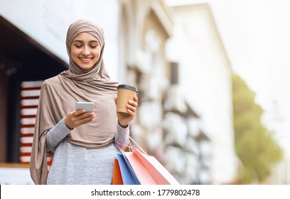 Cheerful muslim girl with shopping bags and cup of coffee using smartphone, spending weekend at shopping mall - Powered by Shutterstock