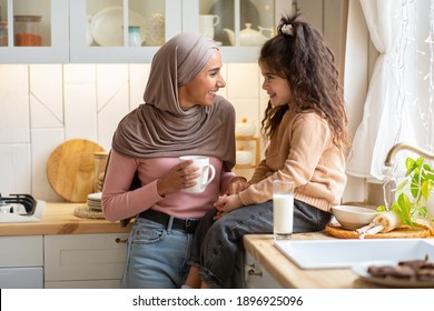 Cheerful muslim mother and her little daughter spending time in kitchen together, having drinks and laughing, having fun at home. Cute child drinking milk and her mom having coffee, free space - Powered by Shutterstock