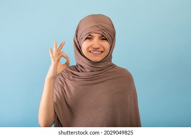 Cheerful Muslim Teen Girl In Hijab Showing Okay Gesture On Blue Studio Background. Positive Arab Adolescent In Islamic Headscarf Recommending Or Approving Something, Smiling At Camera