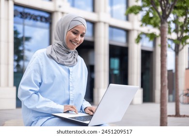 A cheerful Muslim woman in a blue hijab works on her laptop while sitting outside a contemporary building. She is enthusiastic and focused, showcasing diversity and modernity in work environments. - Powered by Shutterstock