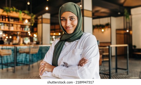 Cheerful Muslim woman looking at the camera while sitting alone in a coffee shop. Young woman with a headscarf sitting in a cafe with her arms crossed. - Powered by Shutterstock