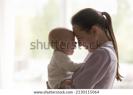 Cheerful new mom having fun with baby, holding kid in arms, hugging with face touches, laughing, enjoying motherhood, maternity leave. Young mother cuddling child with love, tenderness, care