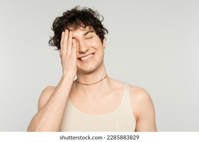 cheerful nonbinary model in tank top and beads covering face with hand and smiling with closed eyes isolated on grey