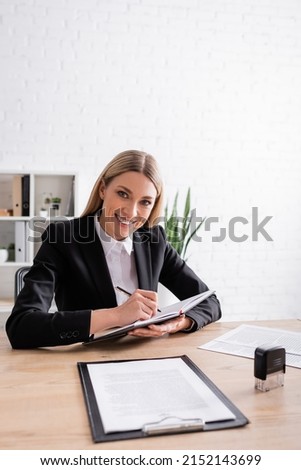 cheerful notary looking at camera while writing in notebook at workplace