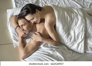 Cheerful nude couple under the white blanket on the bed. Guy holds his hands under the chin. Girl lies on his back. They both smiling. Closeup. Horizontal.
