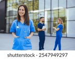 Cheerful nurse in blue scrubs stands outside a modern medical facility, exuding confidence and professionalism. Two colleagues converse in the background, highlighting teamwork in healthcare settings.