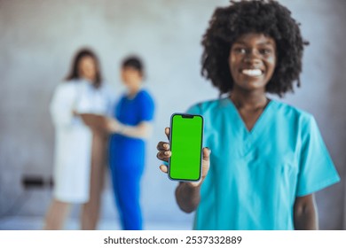 A cheerful nurse in scrubs presents a smartphone with a blank green screen, ideal for medical apps. Blurred healthcare professionals in the background suggest a clinical setting. - Powered by Shutterstock