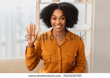 Cheerful at office African American woman with a radiant smile and natural curly hair wearing a mustard blouse waves hello, exuding friendliness and warmth at home office interior