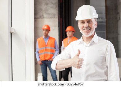 Cheerful Old Architect Is Giving His Thumbs Up. His Project Was Done Perfectly. The Man Is Smiling And Looking At The Camera Happily. Two Builders Are Standing Behind Him And Relaxing