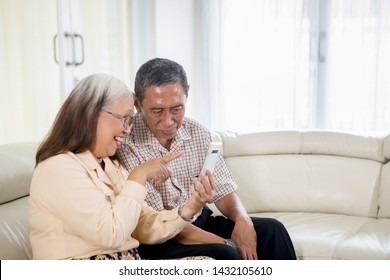 Cheerful Old Couple Making A Video Call By Using A Smartphone At Home While Sitting On The Sofa