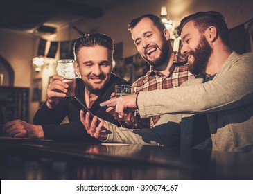 Cheerful Old Friends Having Fun With Smartphone And Drinking Draft Beer At Bar Counter In Pub.