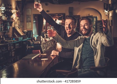 Cheerful Old Friends Having Fun Watching A Football Game On TV And Drinking Draft Beer At Bar Counter In Pub.