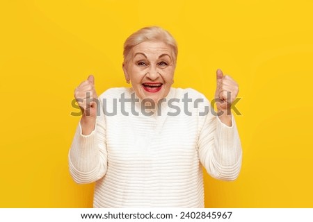 cheerful old grandmother in white sweater rejoices in victory and luck on yellow isolated background, elderly woman in wrinkles celebrates success and wins