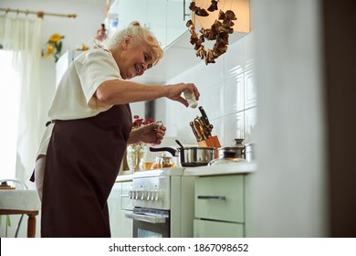 Cheerful Old Lady Adding Salt To Soup And Smiling While Standing By The Stove With Cooking Pots