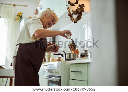 Cheerful old lady adding salt to soup and smiling while standing by the stove with cooking pots