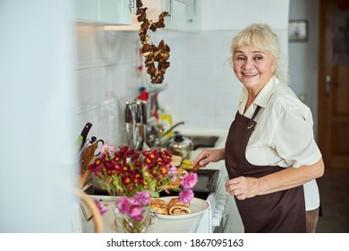 Cheerful Old Lady In Apron Holding Piece Of Pie And Smiling While Cooking Dinner At Home