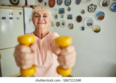 Cheerful Old Lady Holding Yellow Dumbbells And Smiling While Having Morning Workout In Kitchen