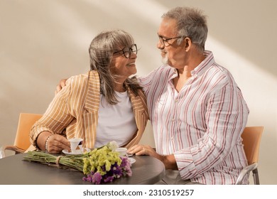 Cheerful old man hugging woman in casual sit at table, enjoy coffee together in cafe outdoor. Love, relationships, free time and weekend, retirement and romantic date at summer - Shutterstock ID 2310519257