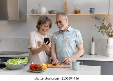 Cheerful old mature retired woman showing funny photo video content or online recipe on cellphone to smiling middle aged husband, enjoying cooking healthy food or preparing meal in modern kitchen. - Powered by Shutterstock