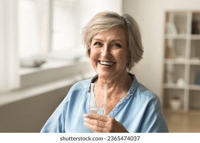 Cheerful old mature woman holding clear glass of fresh mineral water, promoting hydration for elderly skincare, healthy metabolism, healthcare, looking at camera with beautiful toothy smile - Powered by Shutterstock