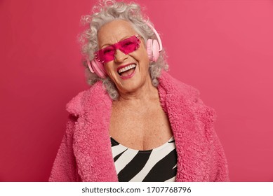 Cheerful Old Woman With Upbeat Mood, Wrinkled Skin, Has Fun On Pension, Listens Music In Modern Stereo Headphones, Wears Trendy Sunglasses, Isolated On Pink Background. Retirement Life Concept