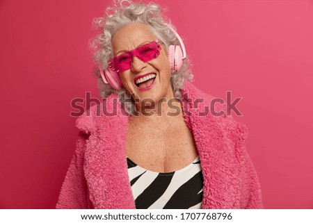 Cheerful old woman with upbeat mood, wrinkled skin, has fun on pension, listens music in modern stereo headphones, wears trendy sunglasses, isolated on pink background. Retirement life concept