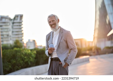 Cheerful older mature adult professional business man, happy senior old businessman wearing suit holding smartphone using mobile cell phone standing outdoor in big city downtown office district. - Shutterstock ID 2231394719