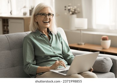 Cheerful older mature freelance business woman in glasses working at home, using laptop, resting on couch, typing on computer, smiling, laughing, looking away. Senior lady shopping online - Powered by Shutterstock