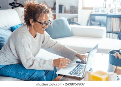 Cheerful older mature freelance business woman in glasses working at home, using laptop, resting on couch, typing on computer, smiling, laughing, looking away. Senior lady shopping online - Powered by Shutterstock