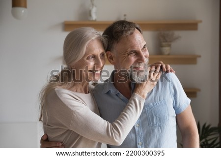 Cheerful older mature married couple hugging at home with love, care, support, tenderness, standing indoors, smiling, laughing, looking away, thinking, dreaming. Retirement, relationship