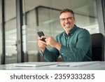 Cheerful older professional businessman, happy middle aged business man entrepreneur laughing looking at camera holding smartphone using cell phone mobile technology sitting at work desk in office.