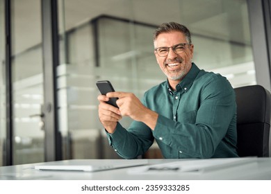 Cheerful older professional businessman, happy middle aged business man entrepreneur laughing looking at camera holding smartphone using cell phone mobile technology sitting at work desk in office. - Powered by Shutterstock