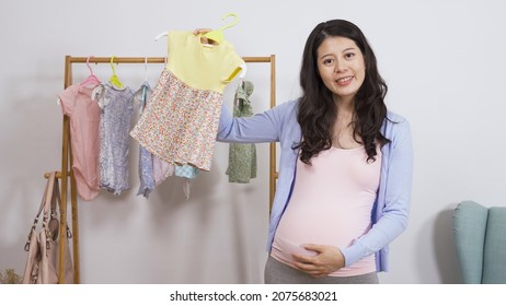 cheerful online mother influencer standing by a rack of baby clothing and looking at camera is recommending the girl dress at white wall background.