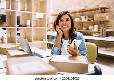 Cheerful online store owner looking away thoughtfully while holding a smartphone. Female entrepreneur preparing orders for shipping in a warehouse. Businesswoman running an e-commerce small business.