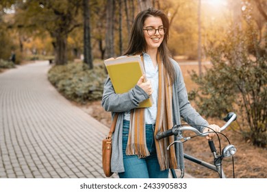 Cheerful optimistic female student holding her college books while walking with bike in public park.