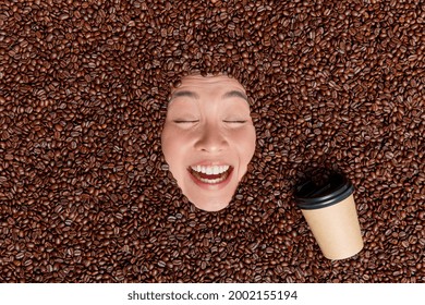 Cheerful optimistic woman surrounded with many coffee beans around drinks espresso from paper disposable cup keeps eyes closed smiles broadly enjoys pleasant aroma or scent. Caffeine addiction