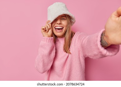 Cheerful optimistic woman wears white panama and jumper has positive playful expression poses for making selfie stretches arm forward camera isolated over pink background takes photo of herself