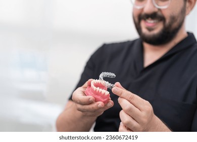 Cheerful orthodontist putting transparent aligners on artificial lower jaw in modern stomatology clinic bearded dentist presenting device for teeth straightening