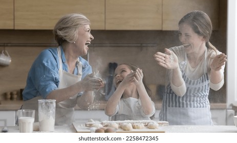 Cheerful overjoyed kid, mother and grandma having fun in kitchen, clapping floury hands over table with bakery food ingredients dough, making cloud, messy, shouting, laughing - Powered by Shutterstock