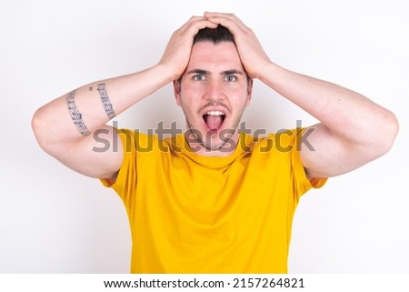 Cheerful overjoyed Young caucasian man wearing yellow t-shirt over white background reacts rising hands over head after receiving great news.