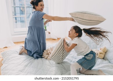 Cheerful and overjoyed young pretty Caucasian mother and her little cute elementary school daughter playing in funny childish activity at home, jumping on bed during pillow fight in bedroom