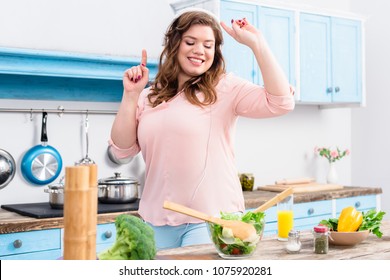 Cheerful Overweight Woman Listening Music In Headphones And Dancing At Table With Fresh Vegetables In Kitchen At Home