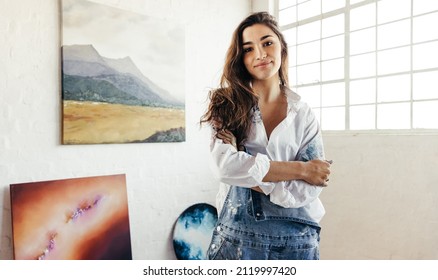 Cheerful painter standing in front of her paintings. Female artist looking at the camera while standing with her arms crossed. Creative young woman displaying a collection of her artwork in her studio