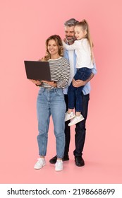 Cheerful Parents And Kid Daughter Using Laptop Browsing Internet Together Over Pink Studio Background. Family Advertising Website Holding Computer. Vertical Shot