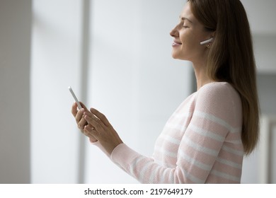 Cheerful Peaceful Pretty Woman With Wireless Headphone In Ear Enjoying Relaxing Music With Closed Eyes, Holding Smartphone, Practicing Stress Relief Meditation Techniques. Side View